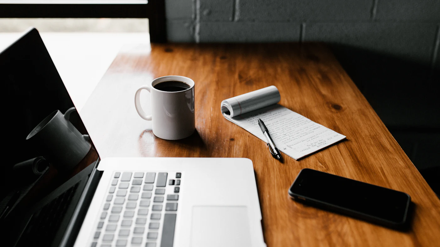 A laptop, a notepad with a pen and a cup of coffee on a wooden desk
